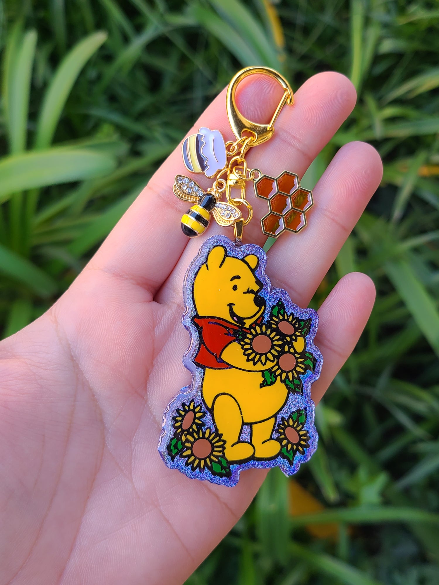 Hand holding a keychain with a cartoon character, bee, and honeycomb against a green leafy background